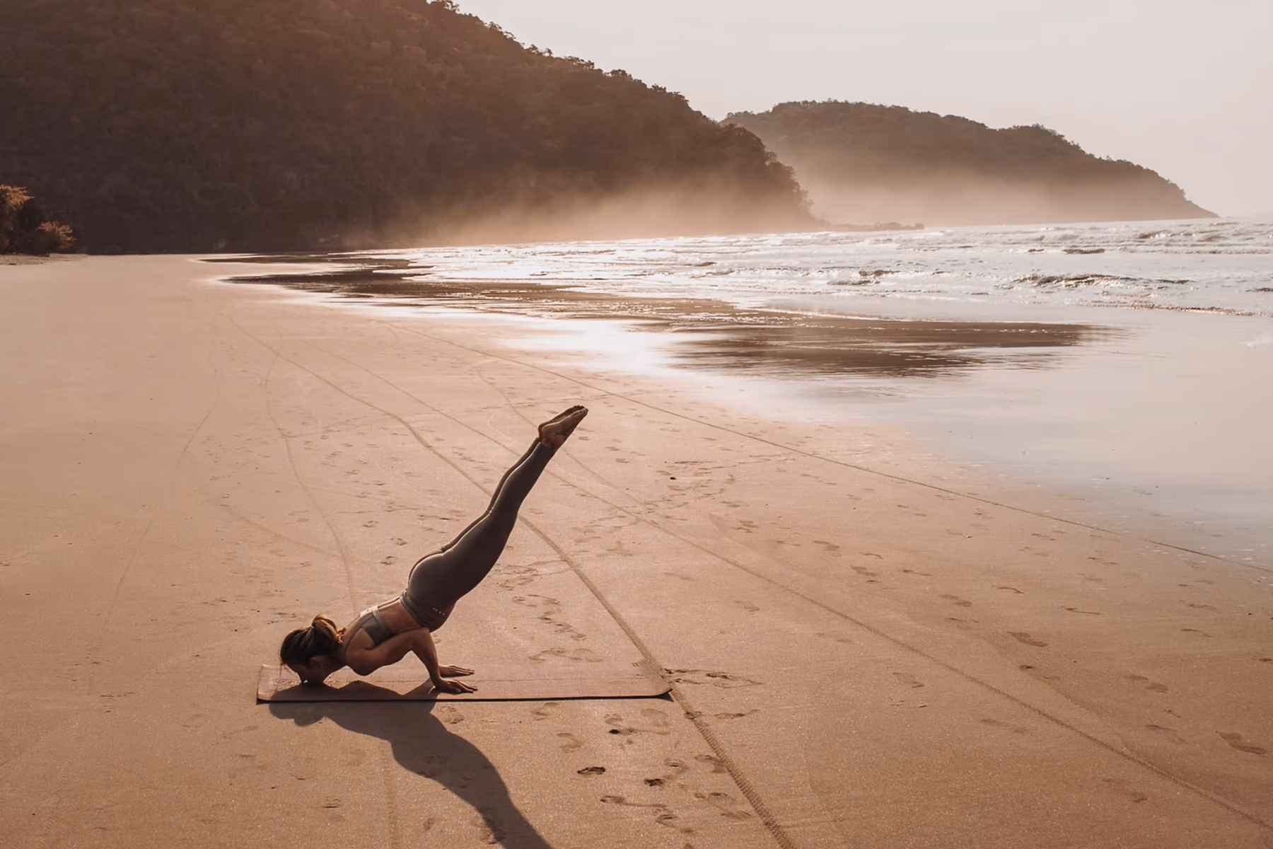 A figure in a forearm-stand yoga pose on an empty golden-hour beach, soft sea mist behind the headland — Bötaniqa's mood of integrated beauty, body, and mind