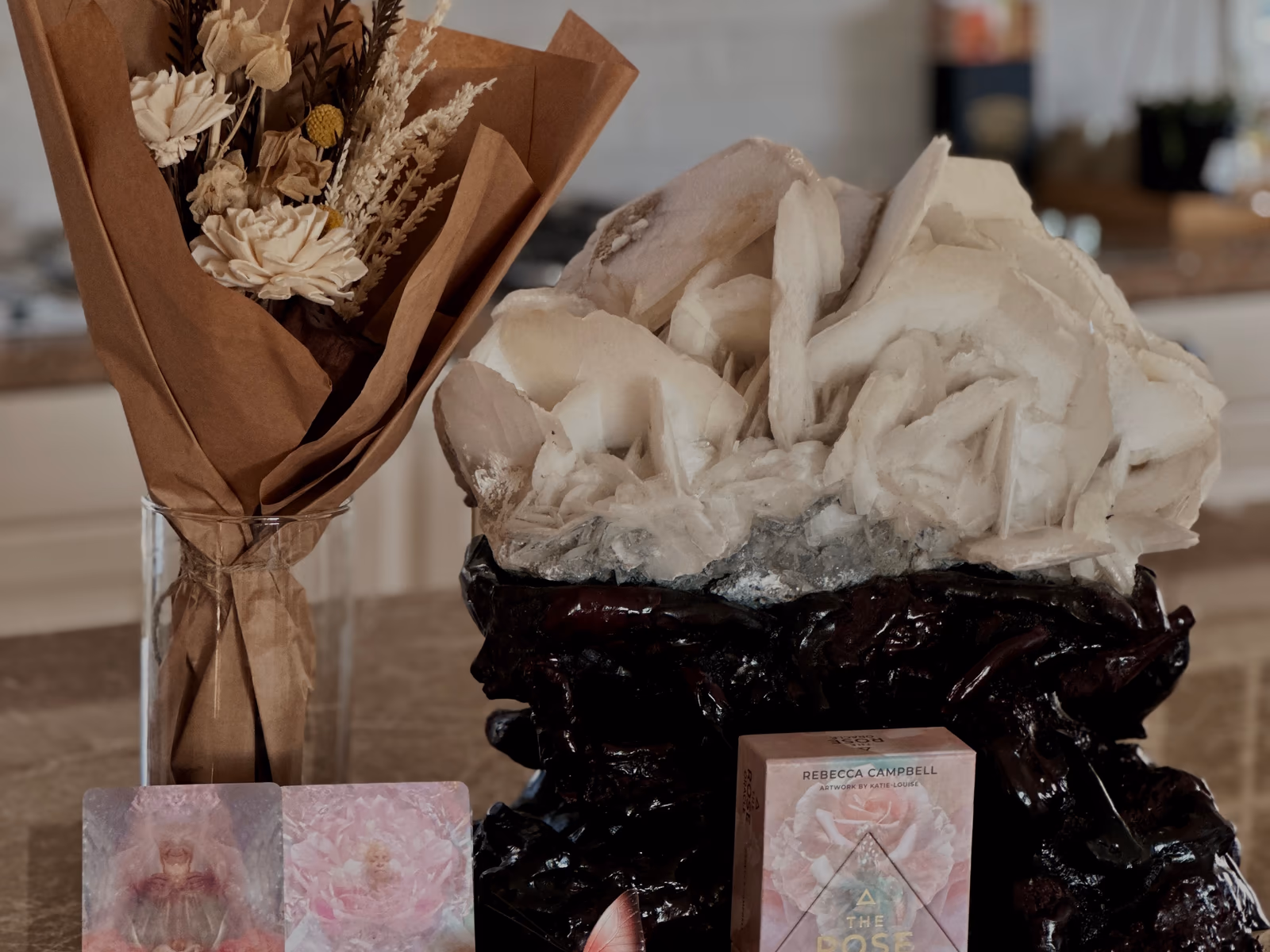 A dried-flower bouquet, a pale crystal cluster, and an oracle deck arranged on a kitchen bench
